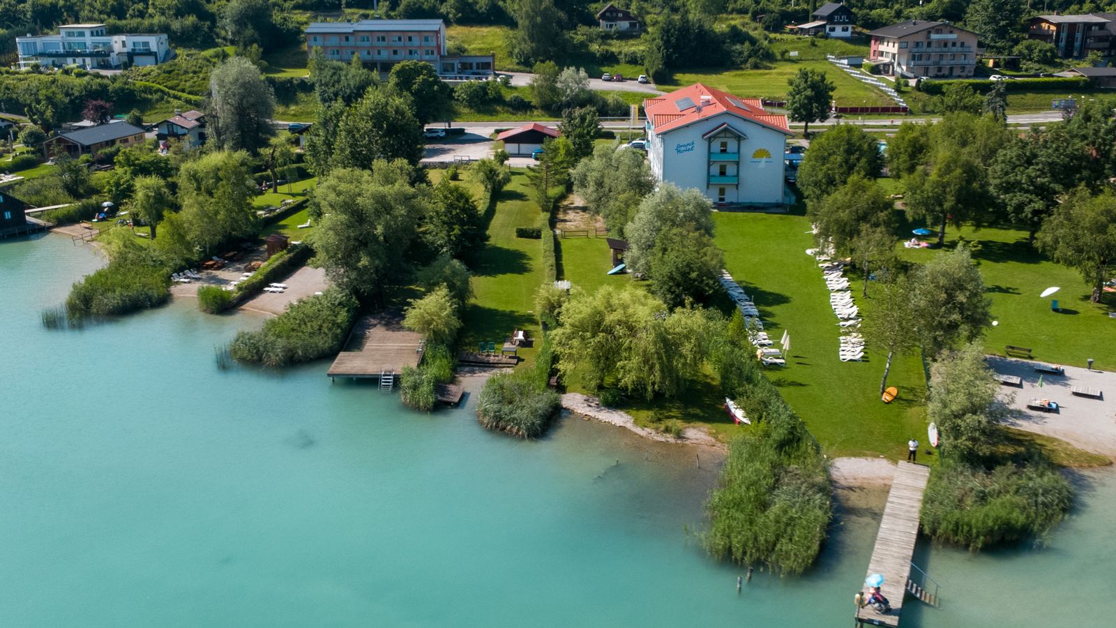 Aerial view of Strandhotel Faak with its private beach, lawn and the turquoise water of Lake Faak